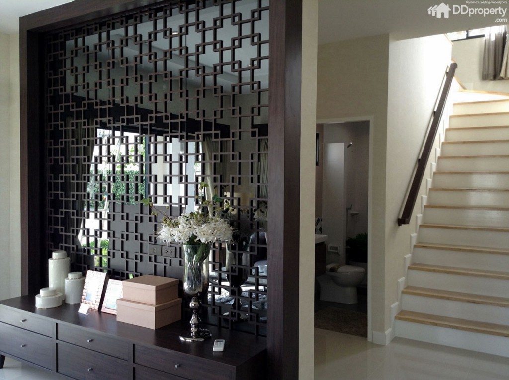 กลับมาดูฝั่งตรงข้ามมุมรับแขก ทางโครงการตกแต่งผนังกั้นแบ่งพื้นที่ด้วยไม้แกะสลักติดกระจกแบบจีน มีลิ้นชักเก็บของไว้เป็นไอเดีย  ซึ่งด้านหลังเป็นประตูทางเข้าจากโรงจอดรถ มีห้องน้ำอยู่ใต้บันได