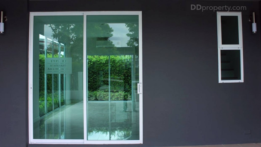 พื้นที่หน้าบ้านเป็นกำแพงคอนกรีตฉาบเรียบทาด้วยสีเทาเข้ม โทนขรึม ตัดกับขอบประตู และหน้าต่างอะลูมิเนียมสีขาว กระจกเป็นแบบใสเขียวตัดแสง พร้อมโคมไฟติดผนังสองดวง 