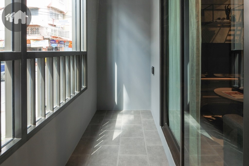 ห้องนั่งเล่นเชื่อมต่อกับระเบียงด้วยประตูกระจกบานเลื่อนแบบสลับ บานใหญ่ พื้นปูกระเบื้องขนาด 30x30 เซนติเมตร ระเบียงด้านข้างกั้นเป็นกำแพงทึบป้องกันฝนสาดได้ รั้วระเบียงสูงประมาณ 1.10 เมตร