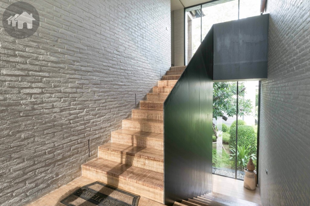 โถงบันไดจากทางเชื่อมที่จอดรถ ซึ่งอยู่ระหว่างกลางของชั้นล่างกับชั้นบน ผนังอิฐโชว์แนว พื้นไม้โอ๊คขัดลาย และราวกันตกใช้แผ่นเหล็กทึบสีดำ กระจกเทมเปอร์บานใหญ่ เปิดรับวิวสวน