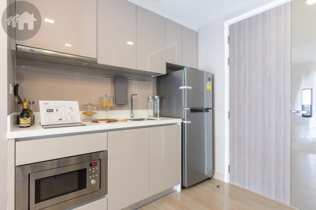 The kitchen is outfitted with high-gloss upper and lower cabinets, extended around 60 cm. from the wall and spread around 1.55 metres in length. There’s a space left for a refrigerator on the right. The countertop is artificial stone in sleek white colour, contrasting with light brown backsplash on the upright panel